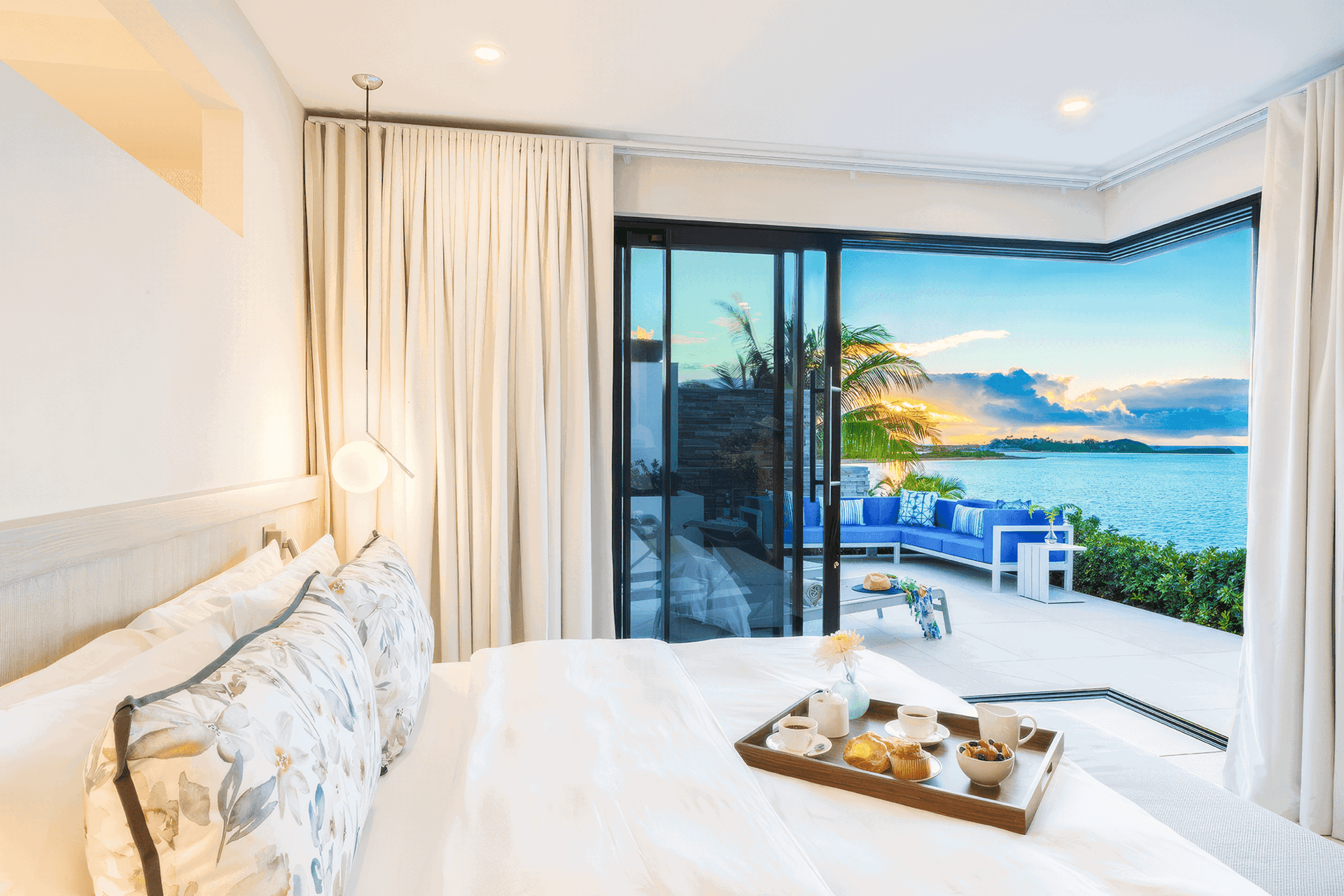 Luxury bedroom with folding sliding glass doors opening to a waterfront patio overlooking sunset views.